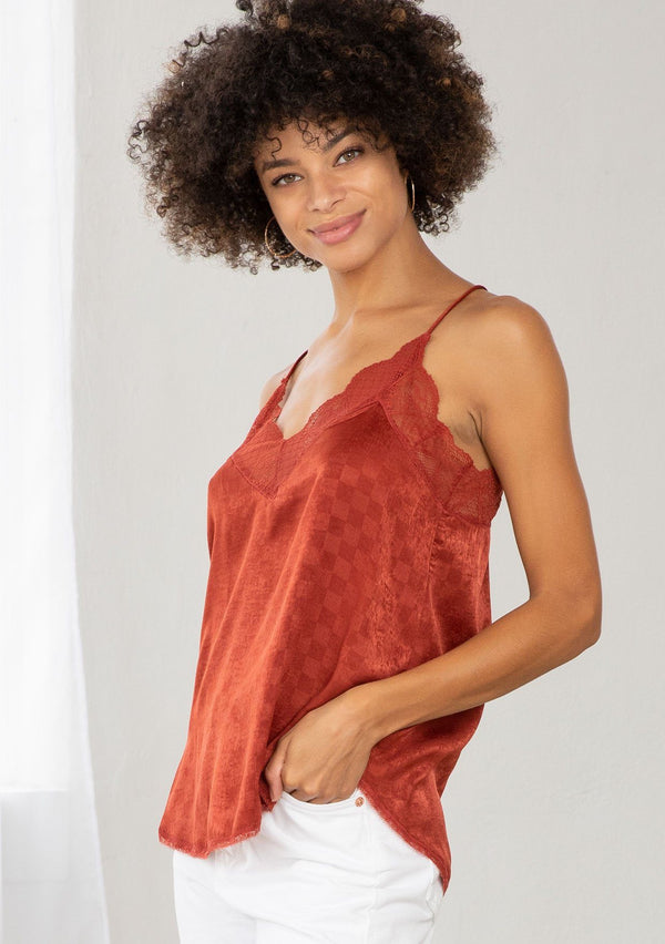 [Color: Burnt Rust] A model wearing a rust red checkered jacquard silky slip camisole. With adjustable spaghetti straps and a lace trim v neckline and racerback. 