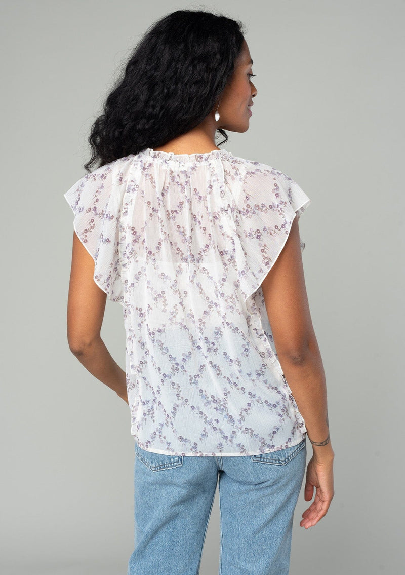 [Color: Ivory/Plum] A back facing image of a brunette model wearing a sheer chiffon bohemian spring top in an ivory white and plum purple floral print. With short flutter sleeves, a split v neckline with ties, and ruffled trim throughout. 