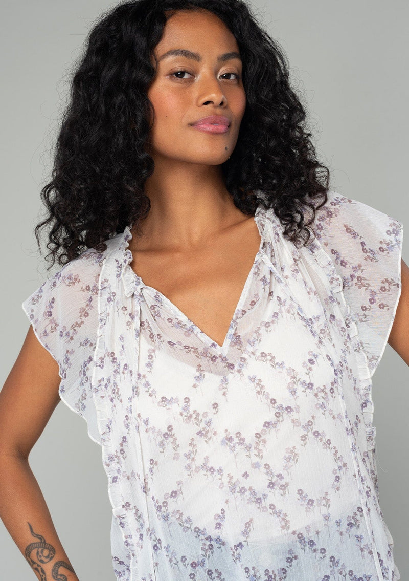 [Color: Ivory/Plum] A close up front facing image of a brunette model wearing a sheer chiffon bohemian spring top in an ivory white and plum purple floral print. With short flutter sleeves, a split v neckline with ties, and ruffled trim throughout. 