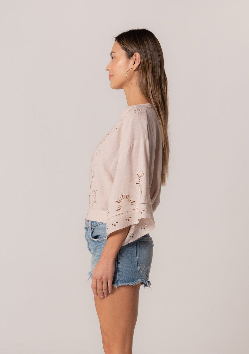 [Color: Natural] A side facing image of a brunette model wearing a bohemian natural light brown embroidered eyelet top with billowy half length sleeves, a button front, and a v neckline.