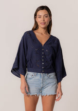 [Color: Navy] A front facing image of a brunette model wearing a bohemian navy blue embroidered eyelet top with billowy half length sleeves, a button front, and a v neckline.