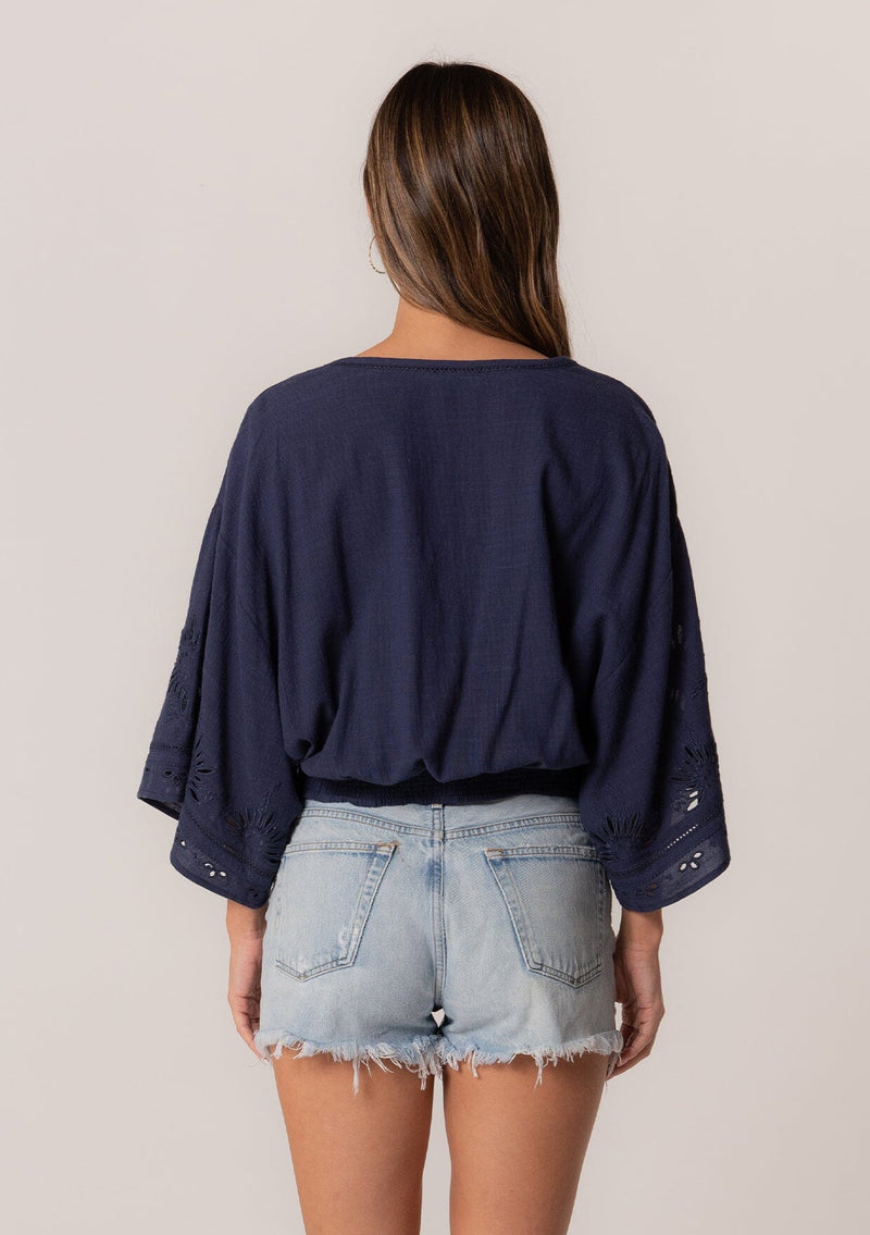 [Color: Navy] A back facing image of a brunette model wearing a bohemian navy blue embroidered eyelet top with billowy half length sleeves, a button front, and a v neckline.