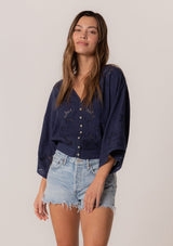 [Color: Navy] A front facing image of a brunette model wearing a bohemian navy blue embroidered eyelet top with billowy half length sleeves, a button front, and a v neckline.
