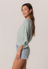 [Color: Seafoam] A side facing image of a brunette model wearing a bohemian seafoam green embroidered eyelet top with billowy half length sleeves, a button front, and a v neckline.
