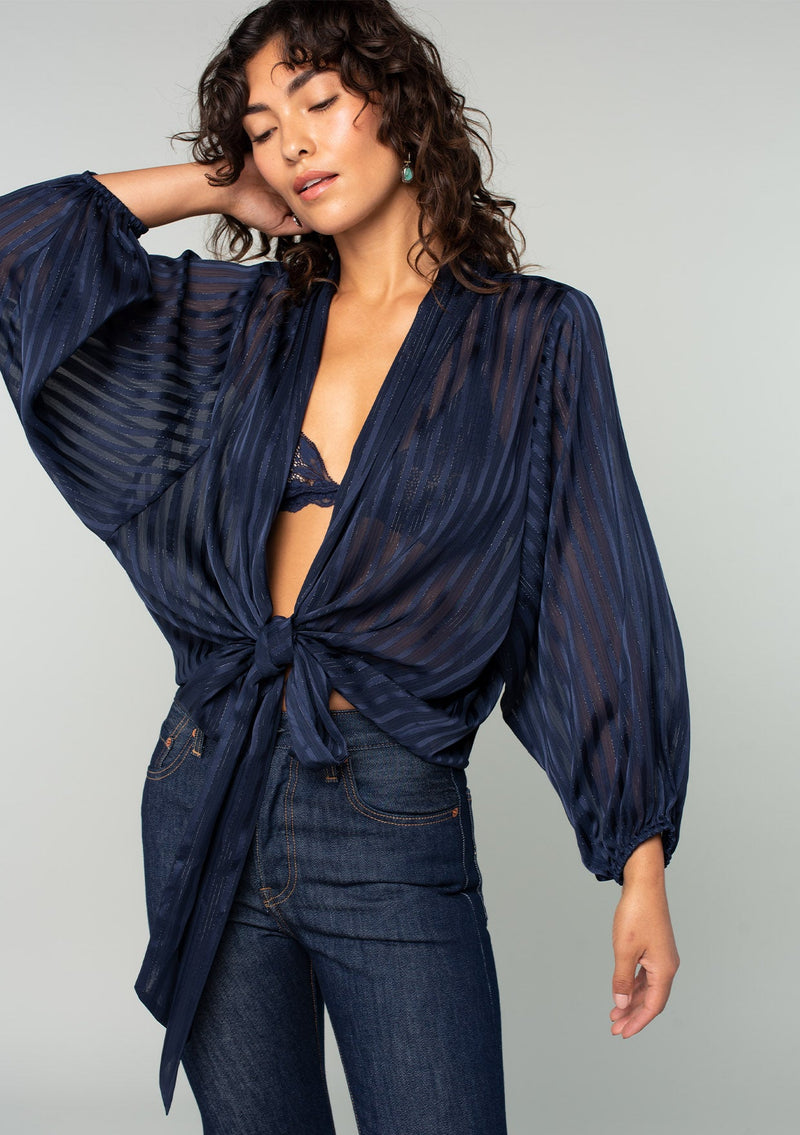 [Color: Navy] A front facing image of a brunette model wearing a sheer navy blue bohemian tie front top in a sparkly lurex stripe. With voluminous long sleeves and a tie front waist that can be styled in multiple ways. 