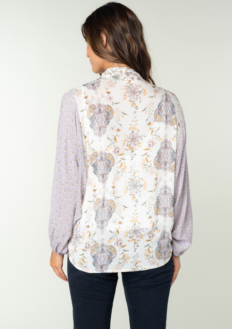 [Color: Natural/Dusty Lilac] A back facing image of a brunette model wearing a lilac purple and natural off white mixed floral print bohemian top. With voluminous long sleeves, a plunging v neckline, and a tie front waist that can be styled in multiple ways. 