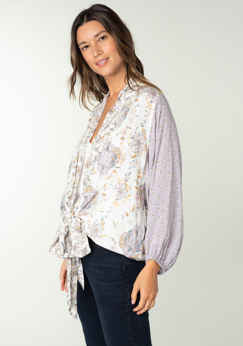 [Color: Natural/Dusty Lilac] A side facing image of a brunette model wearing a lilac purple and natural off white mixed floral print bohemian top. With voluminous long sleeves, a plunging v neckline, and a tie front waist that can be styled in multiple ways. 