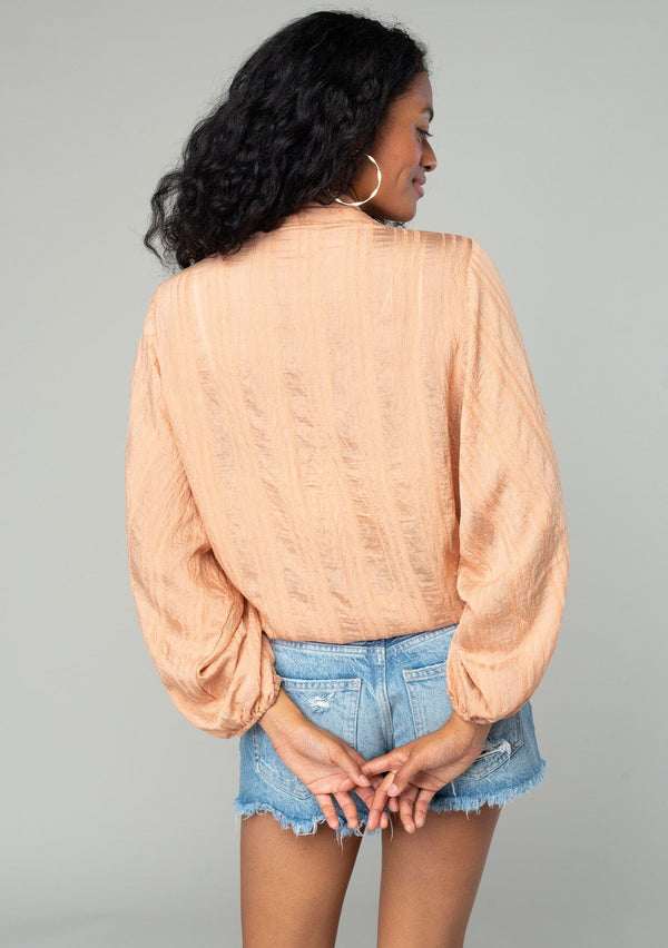[Color: Spice] A back facing image of a brunette model wearing a best selling bohemian tie front resort top in a light orange shadow stripe. With voluminous long sleeves and a tie front detail that can be styled in multiple ways. 
