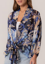 [Color: Navy/Light Blue] A close up front facing image of a brunette model wearing a sheer chiffon tie front top in a blue floral print. With long sleeves, a deep v neckline, a flowy relaxed fit, and a tie front waist detail. 