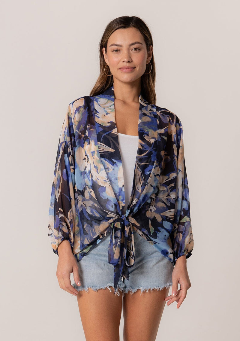 [Color: Navy/Light Blue] A front facing image of a brunette model wearing a sheer chiffon tie front top in a blue floral print. With long sleeves, a deep v neckline, a flowy relaxed fit, and a tie front waist detail. 