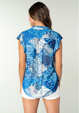 [Color: Blue/Navy] A back facing image of a brunette model wearing a bright blue floral patchwork print bohemian top. With a button front, short flutter sleeves, and neck ties. 