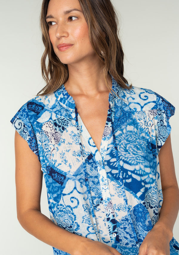 [Color: Blue/Navy] A close up front facing image of a brunette model wearing a bright blue floral patchwork print bohemian top. With a button front, short flutter sleeves, and neck ties. 