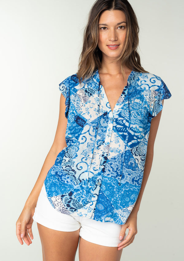 [Color: Blue/Navy] A front facing image of a brunette model wearing a bright blue floral patchwork print bohemian top. With a button front, short flutter sleeves, and neck ties. 