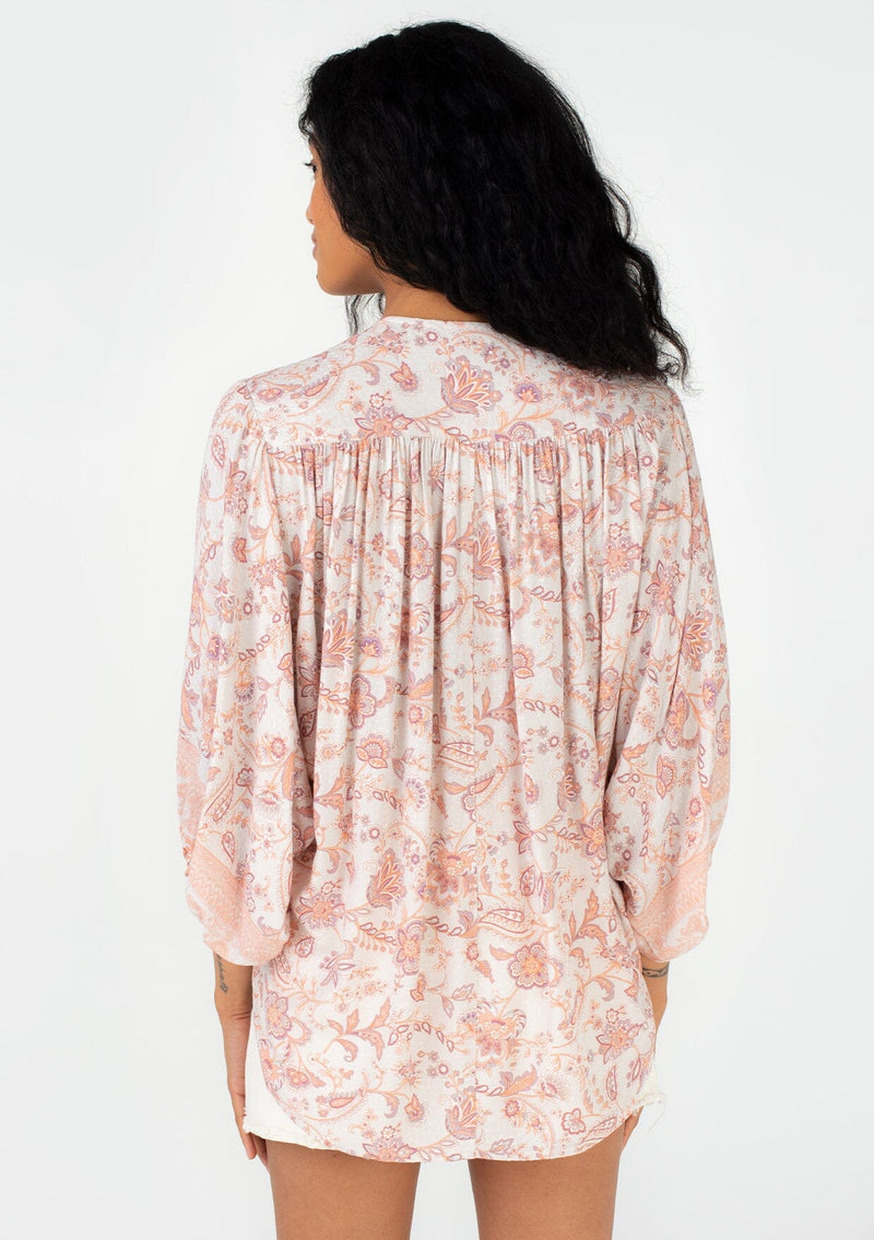 [Color: Natural/Clay] A back facing image of a brunette model wearing a flowy bohemian spring blouse in a pink floral border print. With three quarter length voluminous sleeves, a surplice v neckline, and a high low hemline. 