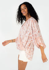 [Color: Natural/Clay] A side facing image of a brunette model wearing a flowy bohemian spring blouse in a pink floral border print. With three quarter length voluminous sleeves, a surplice v neckline, and a high low hemline. 