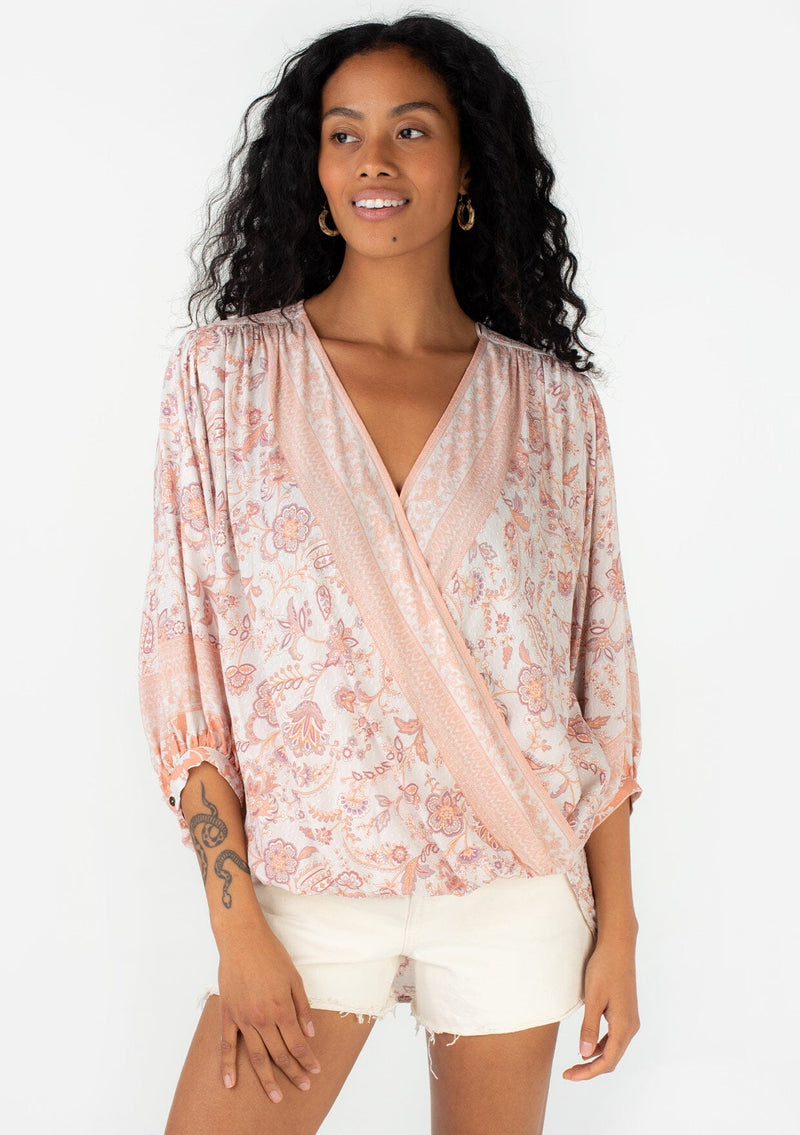 [Color: Natural/Clay] A front facing image of a brunette model wearing a flowy bohemian spring blouse in a pink floral border print. With three quarter length voluminous sleeves, a surplice v neckline, and a high low hemline. 