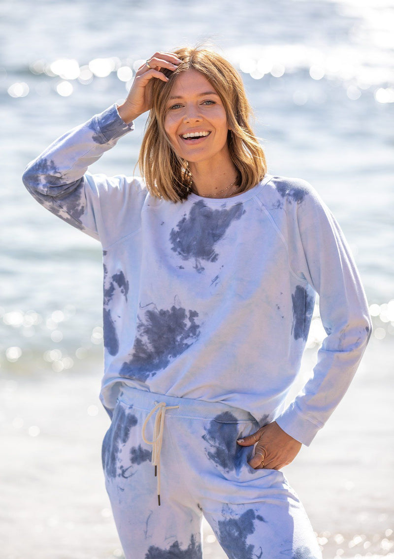 [Color: Ink Combo] A woman standing outside wearing a classic cotton sweatshirt in a splatter tie dye wash. Featuring a crew neckline, long raglan sleeves, and a relaxed fit. 