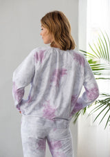[Color: Lavender Combo] A woman standing outside wearing a classic cotton sweatshirt in a splatter tie dye wash. Featuring a crew neckline, long raglan sleeves, and a relaxed fit. 