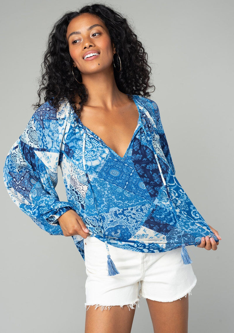 [Color: Blue/Navy] A front facing image of a brunette model wearing a classic flowy bohemian blouse in a blue floral patchwork print. With voluminous long sleeves, a split neckline with tassel ties, and a loose relaxed fit.