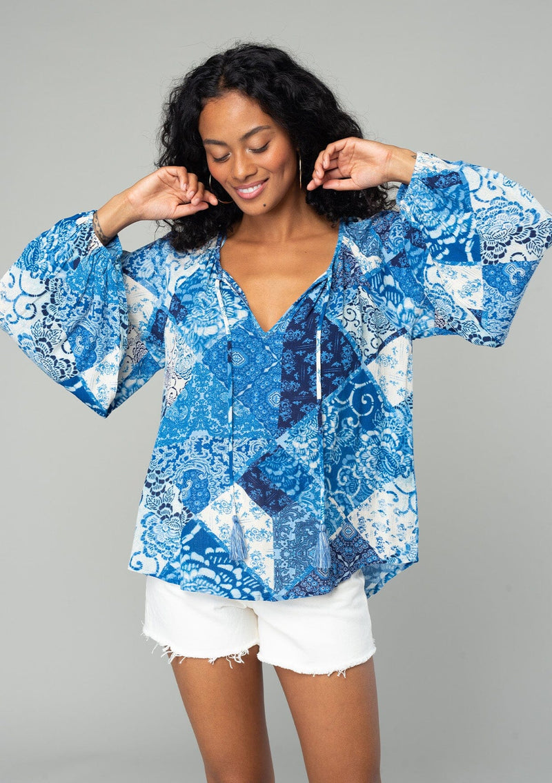 [Color: Blue/Navy] A half body front facing image of a brunette model wearing a classic flowy bohemian blouse in a blue floral patchwork print. With voluminous long sleeves, a split neckline with tassel ties, and a loose relaxed fit.