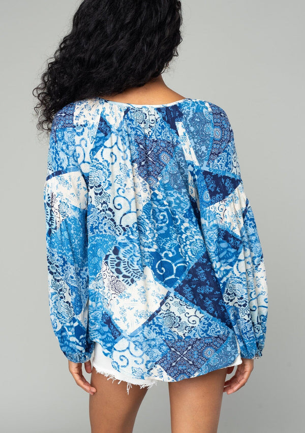 [Color: Blue/Navy] A back facing image of a brunette model wearing a classic flowy bohemian blouse in a blue floral patchwork print. With voluminous long sleeves, a split neckline with tassel ties, and a loose relaxed fit. 