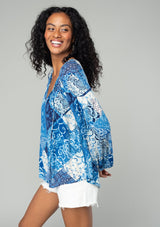 [Color: Blue/Navy] A side facing image of a brunette model wearing a classic flowy bohemian blouse in a blue floral patchwork print. With voluminous long sleeves, a split neckline with tassel ties, and a loose relaxed fit. 
