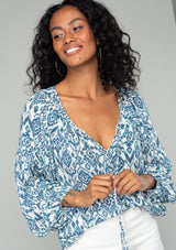 [Color: Ivory/Denim] A close up front facing image of a brunette model wearing a best selling billowy resort blouse in an ivory and blue bohemian diamond print. With exaggerated long sleeves and a split v neckline with tassel ties. 