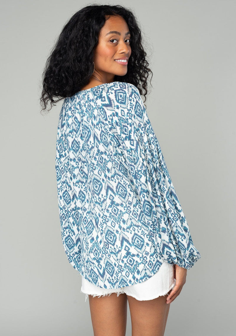 [Color: Ivory/Denim] A back facing image of a brunette model wearing a best selling billowy resort blouse in an ivory and blue bohemian diamond print. With exaggerated long sleeves and a split v neckline with tassel ties. 