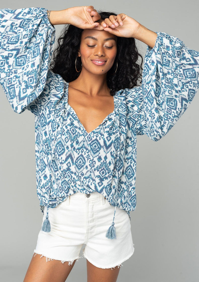 [Color: Ivory/Denim] A half body front facing image of a brunette model wearing a best selling billowy resort blouse in an ivory and blue bohemian diamond print. With exaggerated long sleeves and a split v neckline with tassel ties. 