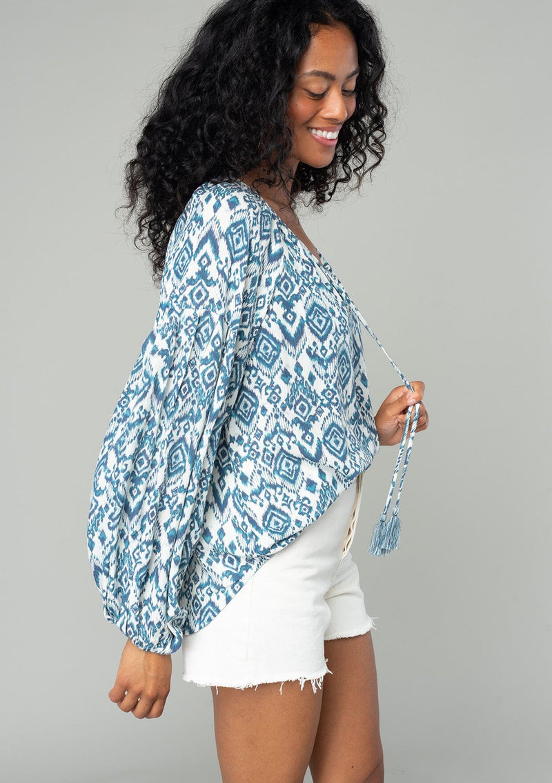 [Color: Ivory/Denim] A side facing image of a brunette model wearing a best selling billowy resort blouse in an ivory and blue bohemian diamond print. With exaggerated long sleeves and a split v neckline with tassel ties. 