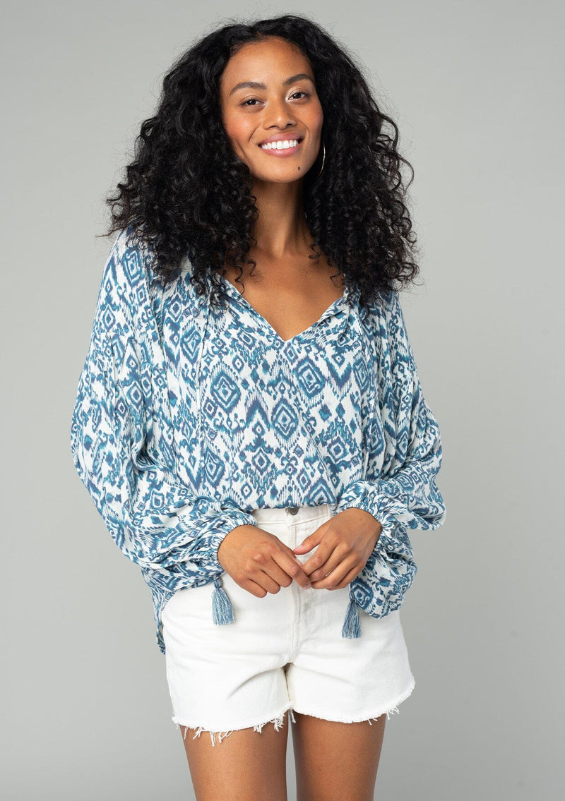 [Color: Ivory/Denim] A front facing image of a brunette model wearing a best selling billowy resort blouse in an ivory and blue bohemian diamond print. With exaggerated long sleeves and a split v neckline with tassel ties. 