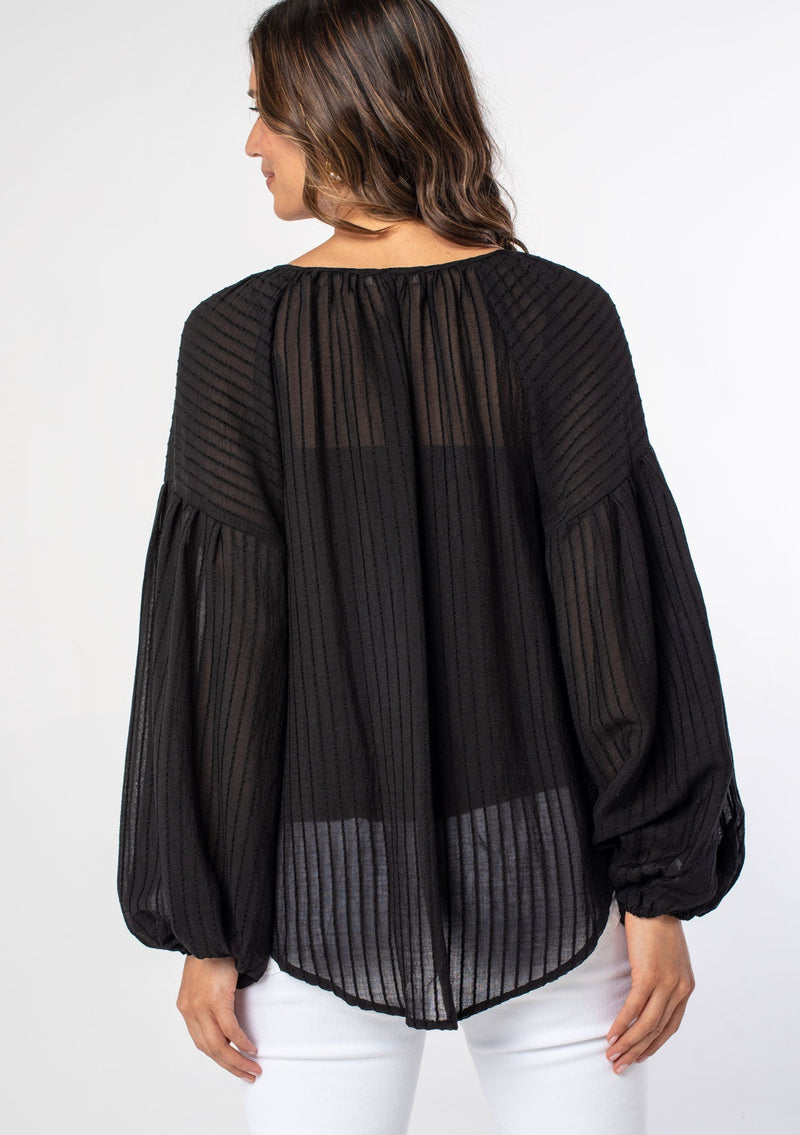 [Color: Black] A woman wearing a black textured shadow striped flowy bohemian peasant top with voluminous long sleeves and a split neckline with tassel ties.