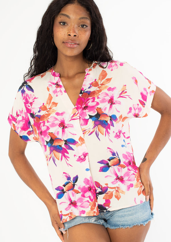 [Color: Ivory/Fuchsia] A front facing image of a black model with long dark wavy hair wearing a white and pink watercolor floral print top with short cuffed sleeves and a v neckline. 