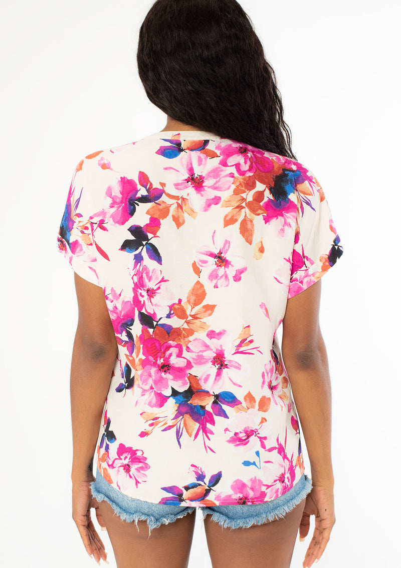 [Color: Ivory/Fuchsia] A back facing image of a black model with long dark wavy hair wearing a white and pink watercolor floral print top with short cuffed sleeves and a v neckline. 