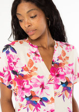 [Color: Ivory/Fuchsia] A close up front facing image of a black model with long dark wavy hair wearing a white and pink watercolor floral print top with short cuffed sleeves and a v neckline. 
