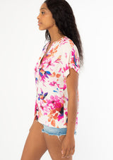 [Color: Ivory/Fuchsia] A side facing image of a black model with long dark wavy hair wearing a white and pink watercolor floral print top with short cuffed sleeves and a v neckline. 