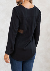 [Color: Black] A model wearing a black blouse with sheer lace inserts. Featuring long sleeves with a button cuff wrist, a self covered button front, and pleated details at the back. 