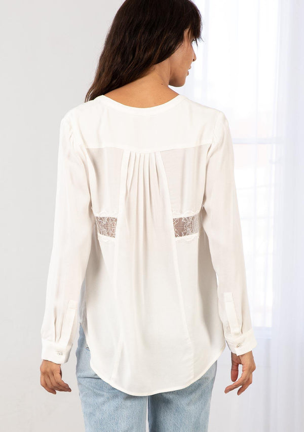 [Color: Ivory] A model wearing an ivory blouse with sheer lace inserts. Featuring long sleeves with a button cuff wrist, a self covered button front, and pleated details at the back. 