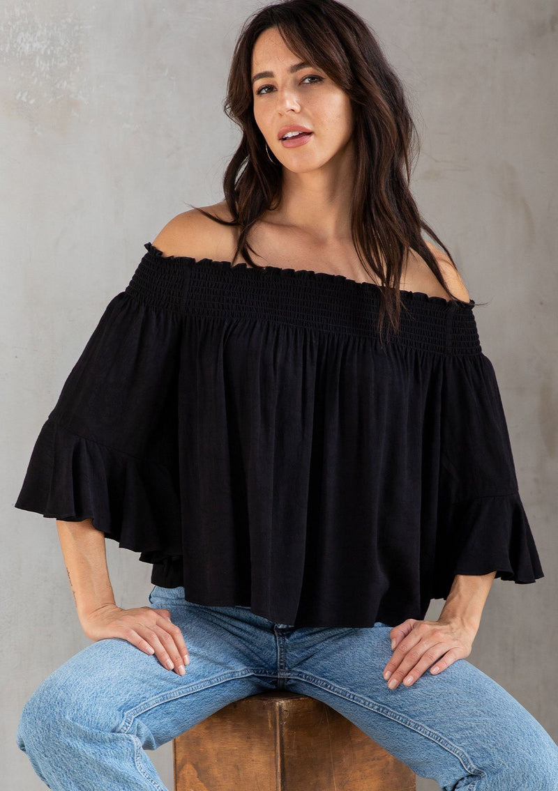 [Color: Black] A model wearing a classic black bohemian blouse in rose jacquard. With three quarter length sleeves, a flounce cuff, and a smocked off shoulder. 