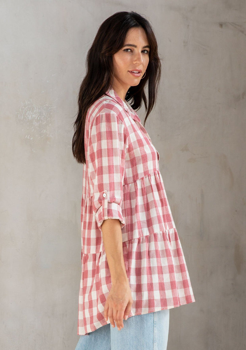 [Color: Dusty Rose/White] A model wearing a relaxed cotton blend pink large gingham button up tunic, with a rolled sleeve, a tiered body, and a long tunic length. 