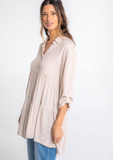 [Color: Tan] A model wearing a relaxed textured checkered jacquard button up tunic, with a rolled sleeve, a tiered body, and a long tunic length.