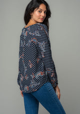 [Color: Charcoal/Dusty Blue] A front facing image of a brunette model wearing a charcoal grey and light blue floral paisley print bohemian blouse. With long sleeves and a v neckline. The model is looking over her shoulder. 