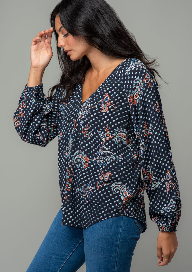 [Color: Charcoal/Dusty Blue] A side facing image of a brunette model wearing a charcoal grey and light blue floral paisley print bohemian blouse. With long sleeves and a v neckline. 