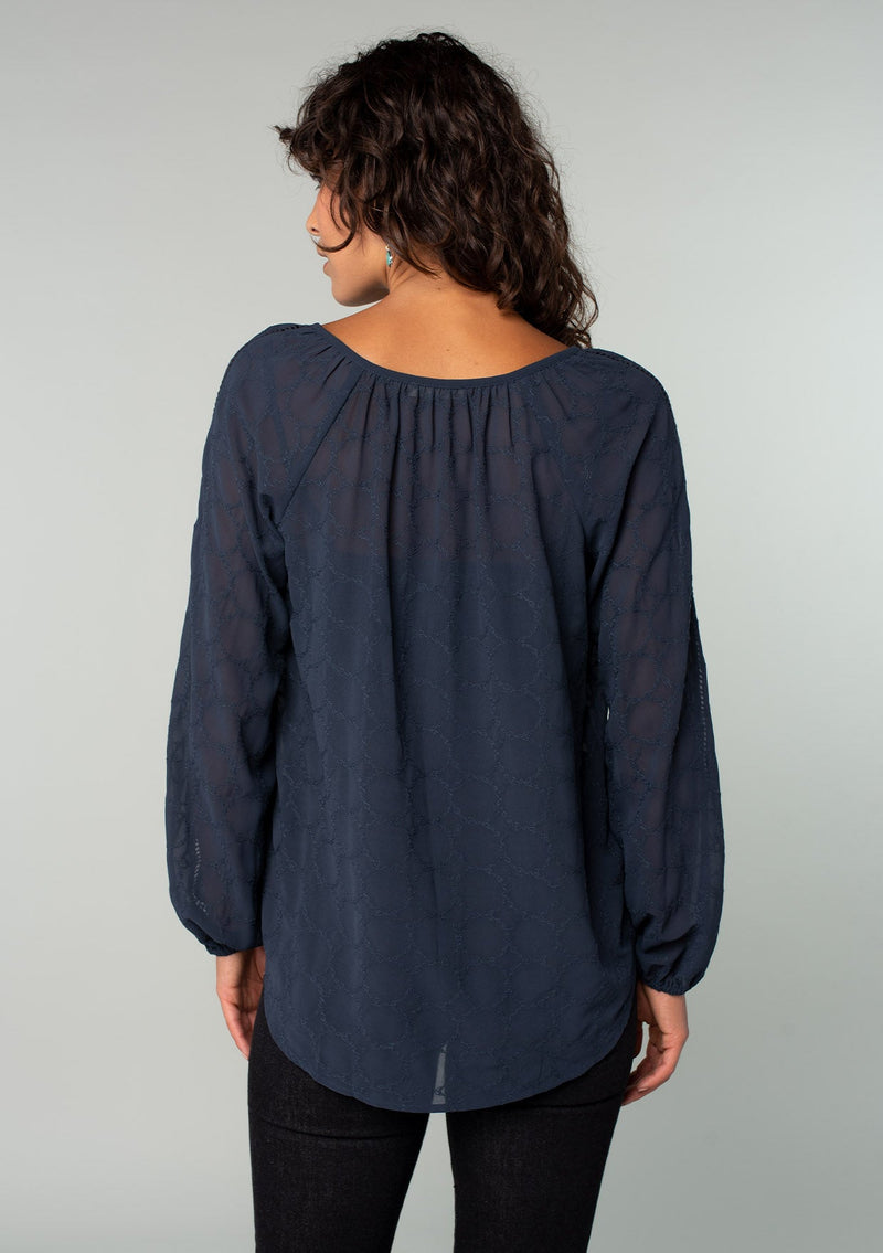 [Color: Ink Blue] A back facing image of a brunette model wearing a navy blue bohemian peasant top in embroidered chiffon. With long sleeves, lattice trim, a v neckline, a button front, and a flowy relaxed fit. 