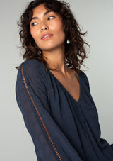 [Color: Ink Blue] A close up side facing image of a brunette model wearing a navy blue bohemian peasant top in embroidered chiffon. With long sleeves, lattice trim, a v neckline, a button front, and a flowy relaxed fit. 