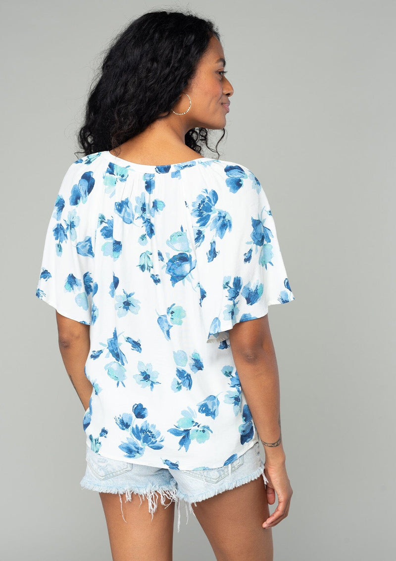 [Color: Ivory/Aqua] A back facing image of a brunette model wearing a classic bohemian spring to in an ivory white and blue floral print top. With short flutter sleeves and a loop button front. 