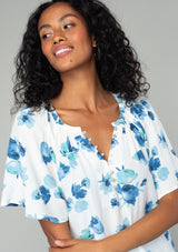 [Color: Ivory/Aqua] A close up front facing image of a brunette model wearing a classic bohemian spring to in an ivory white and blue floral print top. With short flutter sleeves and a loop button front. 