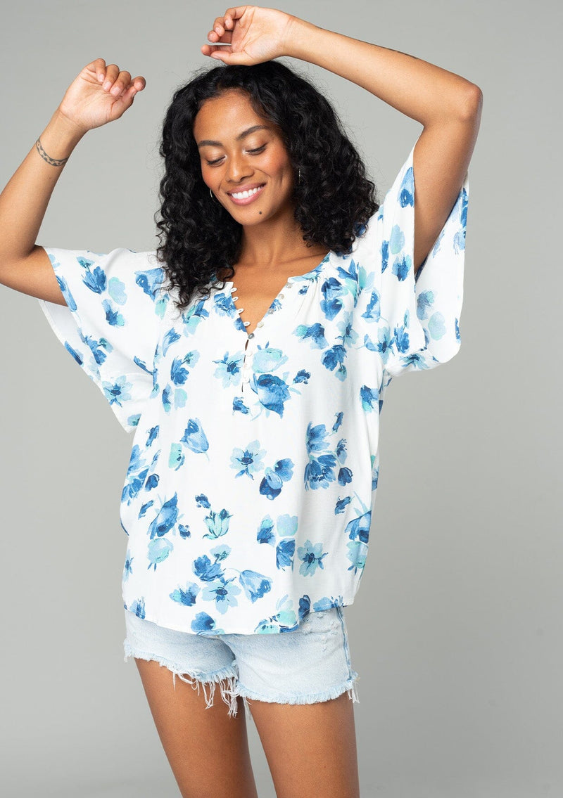 [Color: Ivory/Aqua] A half body front facing image of a brunette model wearing a classic bohemian spring to in an ivory white and blue floral print top. With short flutter sleeves and a loop button front. 