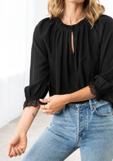 [Color: Black] A model wearing a soft peasant top. With three quarter length voluminous sleeves, a lace wrist cuff, a ruffled neckline with a self covered button loop closure, and a front keyhole detail. 