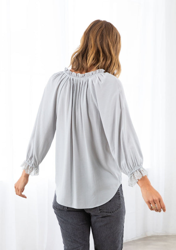 [Color: Sky] A model wearing a soft peasant top. With three quarter length voluminous sleeves, a lace wrist cuff, a ruffled neckline with a self covered button loop closure, and a front keyhole detail. 